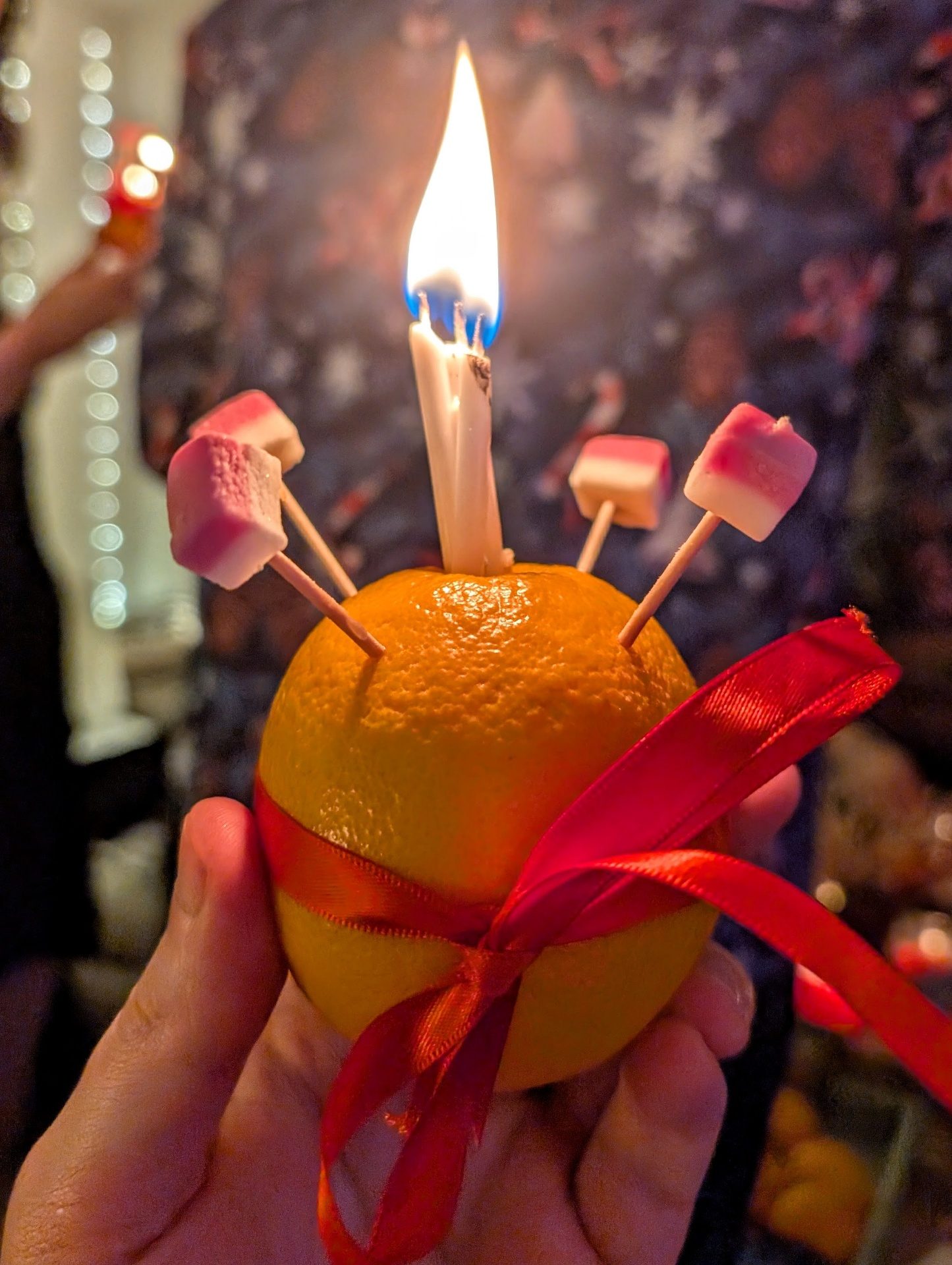 A Christingle orange with lit candle, red ribbon and marshmallow-topped sticks
